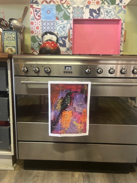 A modern kitchen with a stainless steel oven displaying a colorful abstract painting hanging from the handle. The background features a decorative tiled wall with various patterns.